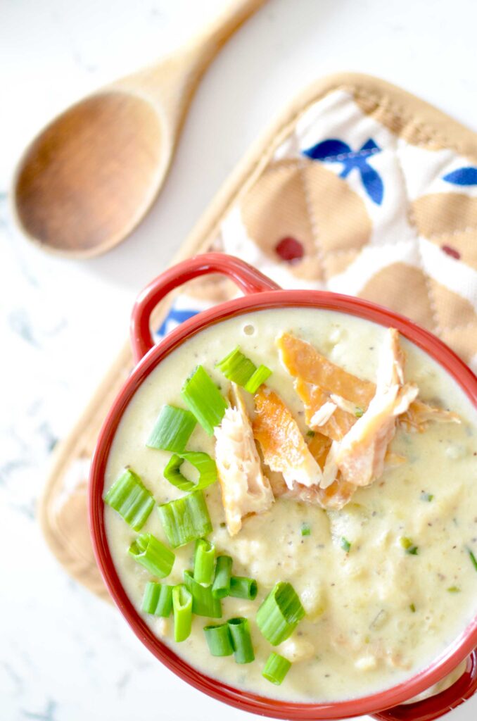 an overhead photo of a red bowl of hearty cullen skink soup. It's a cream colours, with green herbs, and potato chunks. There's extra pieces of smoked white fish pieces added on top and chopped green onions next to it. It's resting on a light brown and cream patterned oven mitt to protect the marble patterned countertops below. There's a wooden spoon lying next to the bowl and mitt on the counter