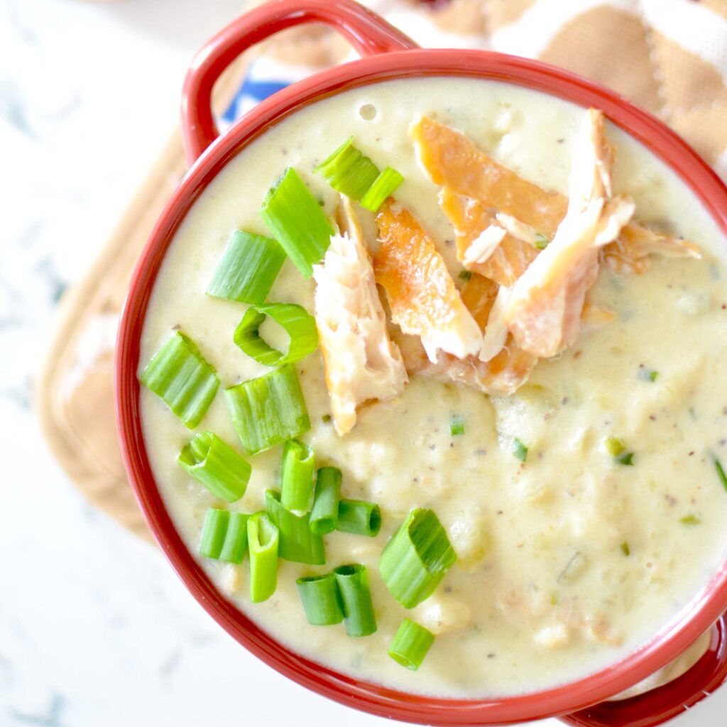 an overhead photo of a red bowl of hearty cullen skink soup. It's a cream colours, with green herbs, and potato chunks. There's extra pieces of smoked white fish pieces added on top and chopped green onions next to it. It's resting on a light brown and cream patterned oven mitt to protect the marble patterned countertops below.