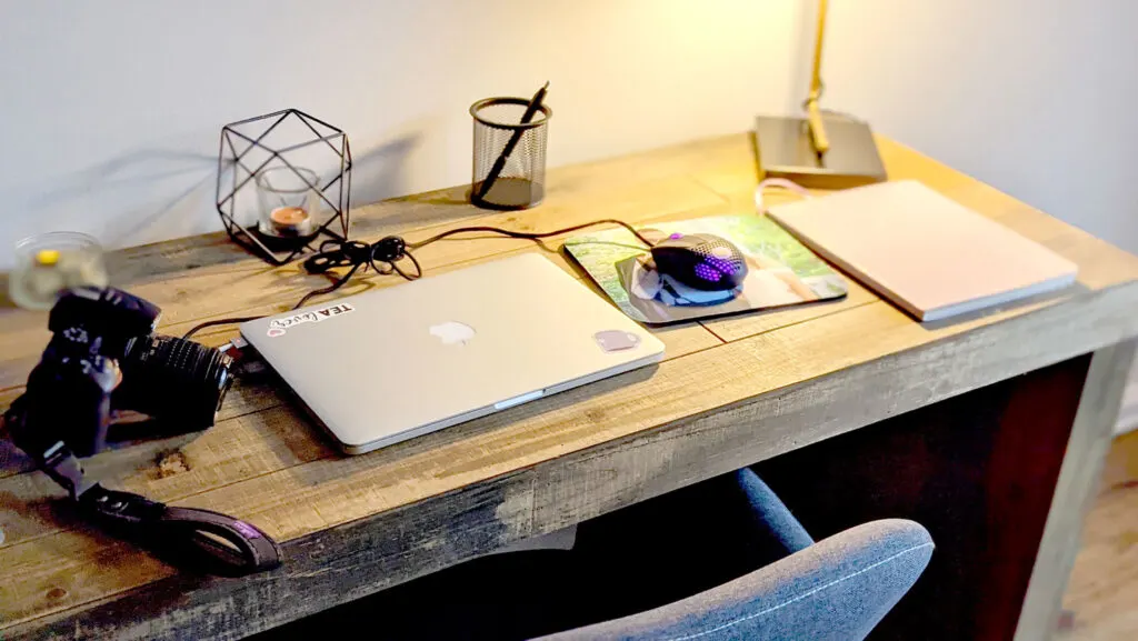 showcasing all the basic office supplies I need on a wood desk with a fabric chair pushed in. There's a DSLR camera, a laptop, mouse and mousepad, and an agenda