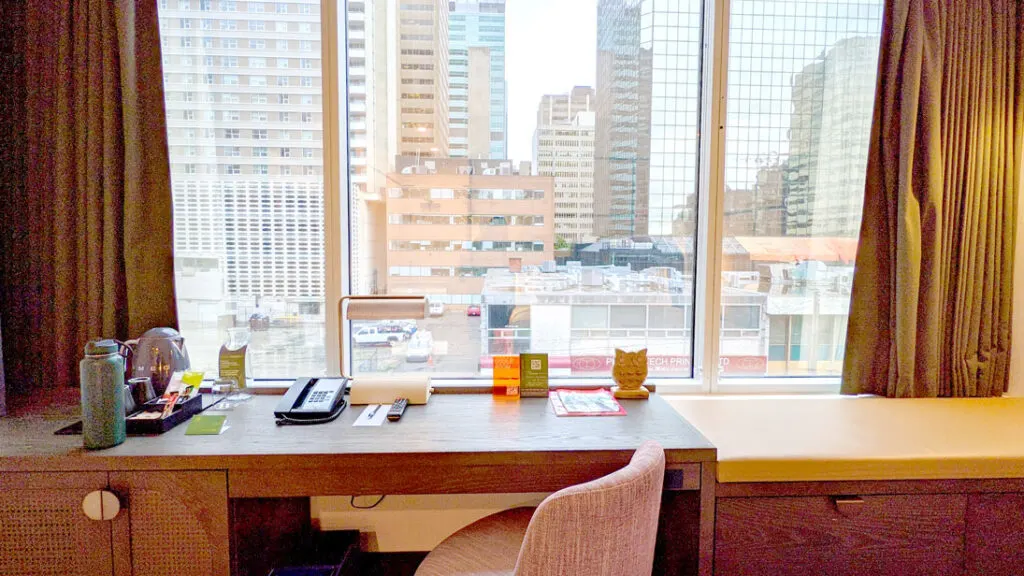 the ultimate work space at a hotel. A fabric swivel chair fits underneath a small wooden desk overlooking a downtown skyline out the window in front of it