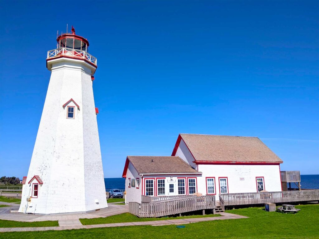 the east point lighthouse Prince Edward island. A tall white lighthouse, with a railing at the top and a small room with 360 degree window all around. There's one window halfway up the lighthouse. The lighthouse also has red paint for the accent colours. Next to the lighthouse is a small white and red house. There's green grass all around, but behind the house and light house you can see the blue water begin