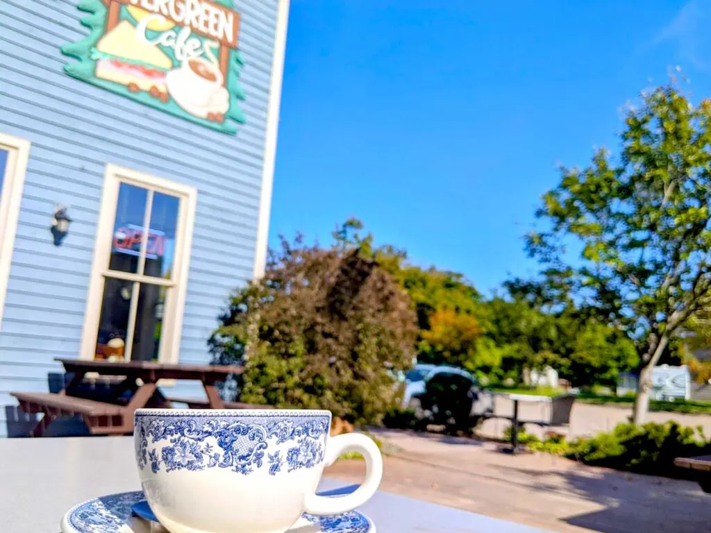 evergreen cafe sourris pei. A victorian decorated white and blue ceramic mug sits on a table outside. There's a picnic table behind it next to a blue building. On top of the building is a large sign that reads: Evergreen Cafe, with a sandwich and coffee.