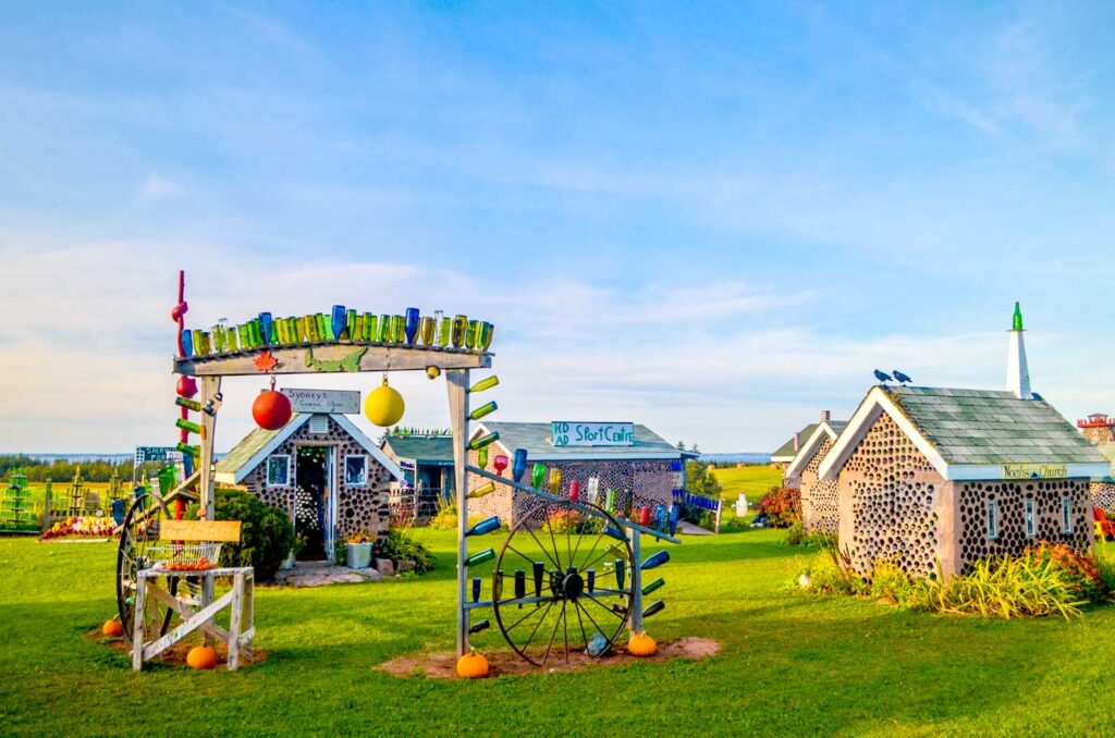 hannahs bottle village in pei during golden hour. There are shadow streaks across the grass, lighting up the concrete and bottle structures. 

There's an archway to walk through, with wood to hold up all different types of bottles on top. Behind the entryway are 4 concrete and glass bottle buildings. 