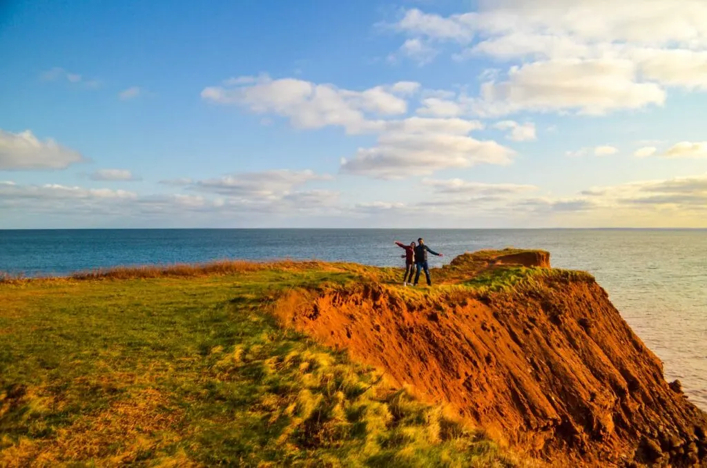 a couple standing in the middle of a red seaside cliff on the north cape coastal drive at edgemont lighthouse. You can tell the sun is starting to set, creating golden hues on the red dirt, and the right side of the image is tinted yellow compared to the left side which is still a very bright blue. 