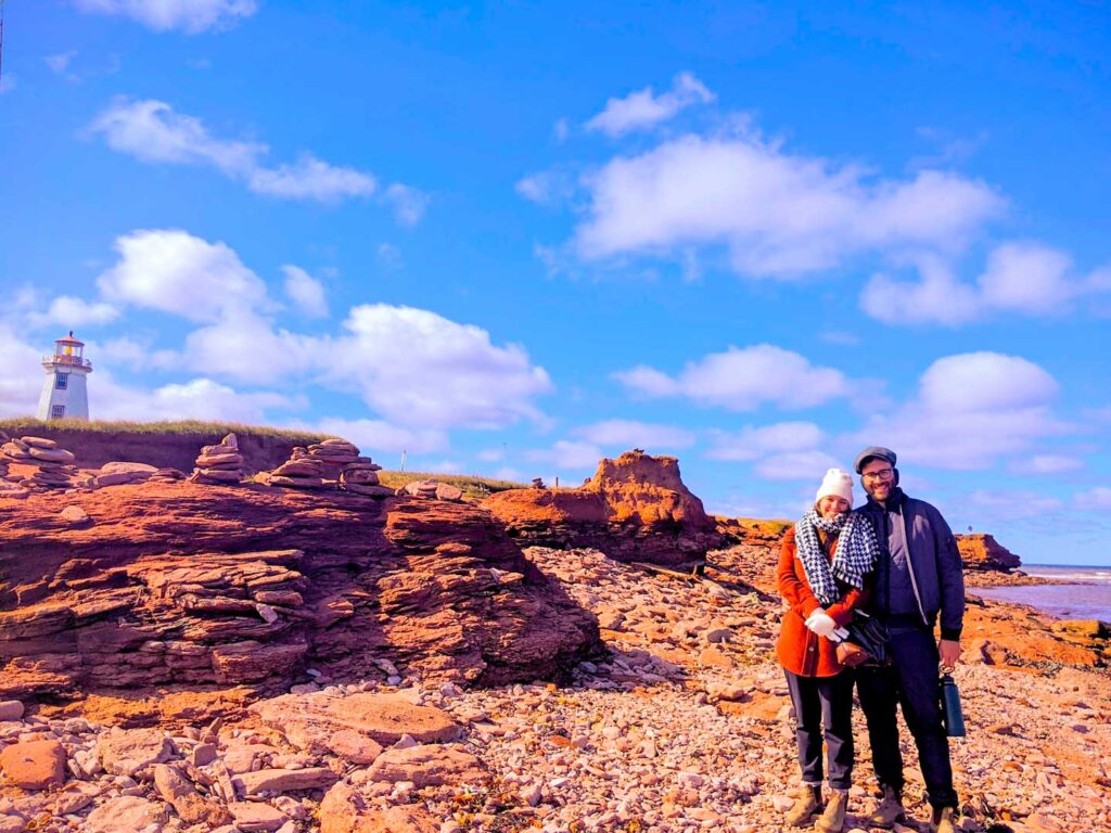 north cape coastal drive lighthouse behind the scales of eroded red rocks on the cliff side during low tide. 

A couple, looking kind of cold, all bundled in jackets, a hat and scarf, on the shore. 