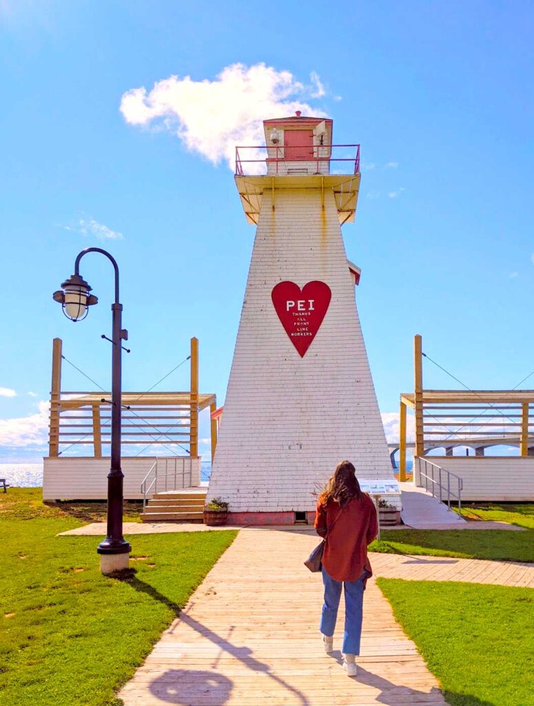 pei central coastal drive lighthouse. A woman in a red wool jacket is walking on a wood side walk towards a white lighthouse that looks like it's seen better days. Yo can tell it's windy because her long brown hair is blowing towards the left. 

 In the middle of the lighthouse is a red heart with PEI written in the middle. 

Behind the lighthouse, you can see the confederation bridge 