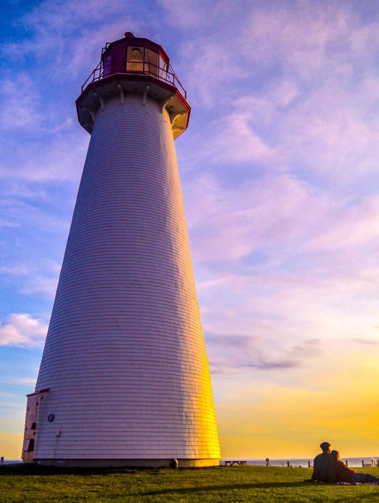 a couple sitting on the grass next to a tall point prim  lighthouse. Yellow shines in from the right side of the photo, lighting up the front of the couple, but creating a long shadow behind them, leading your way straight to the base of the lighthouse. As you go up the lighthouse, the colour changes to pinkish and purplish hues, before turning back into the blue sky behind it. 