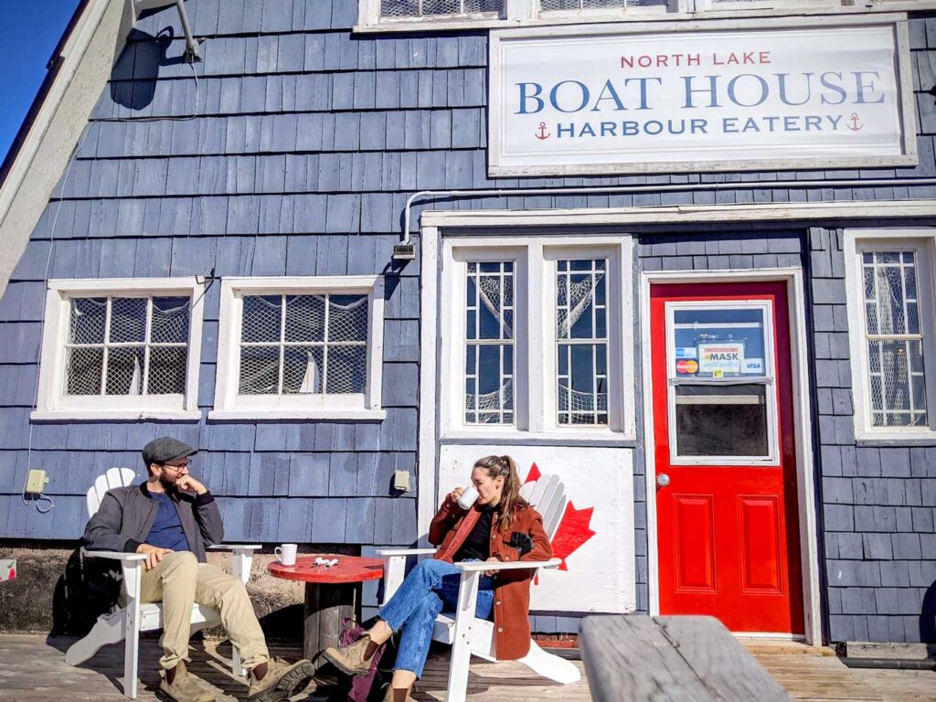 points east coastal drive lunch spot: North Lake Boathouse Harbour Eatery. A couple sits outside on the front patio in Adirondack chairs, in their fall jackets, soaking up the sun enjoying some warm coffee. 

Behind them is a blue shingled building. You can see fishnets over the windows inside. The door is BRIGHT red. Above the door is the sign of the restaurant: North Lake Boathouse Harbour Eatery