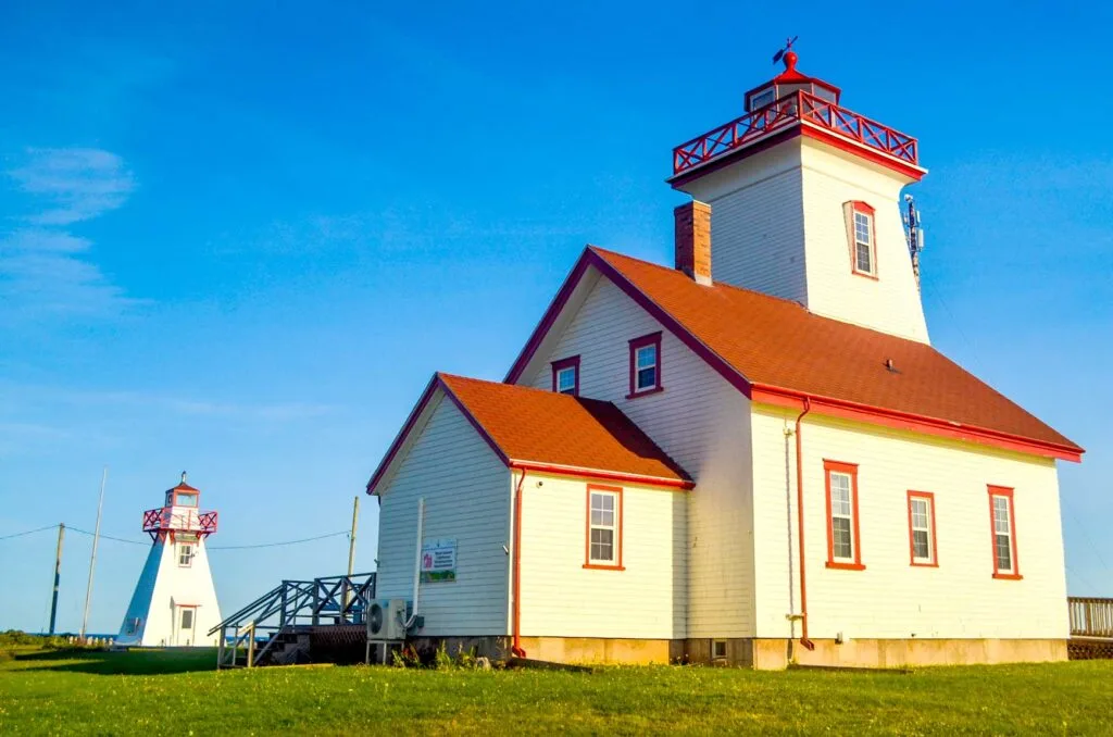 wood islands lighthouse pei. Photographed from a bit below eye level to make the buildings seem taller and take up the photos. You can tell its golden hour because the white paint of the lighthouses has a yellowish tint. 

The building in the foreground looks more like a house, with red roofs, and a lighthouse that sticks up out of it. 

Smaller, in the background, is a more typical lighthouse structure. 