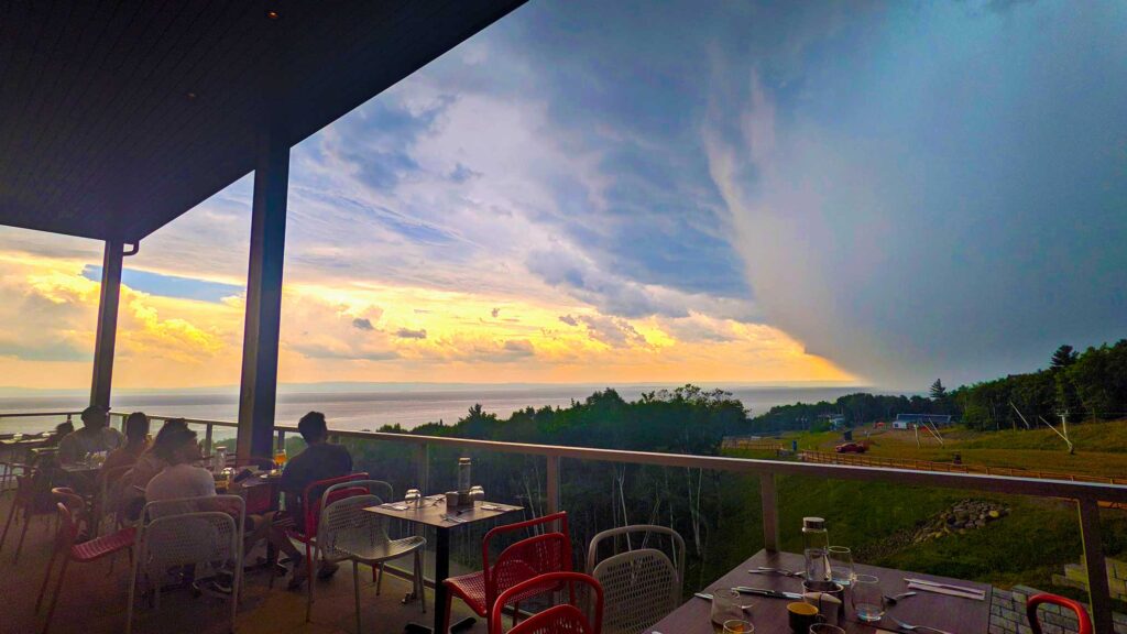 changing-weather-charlevoix-club-med sitting outside on the balcony eating dinner half the sky is a beautiful warm sunset that's being overtaken by an enormous dark grey cloud. other people sitting at the tables are all looking at this at it moves closer