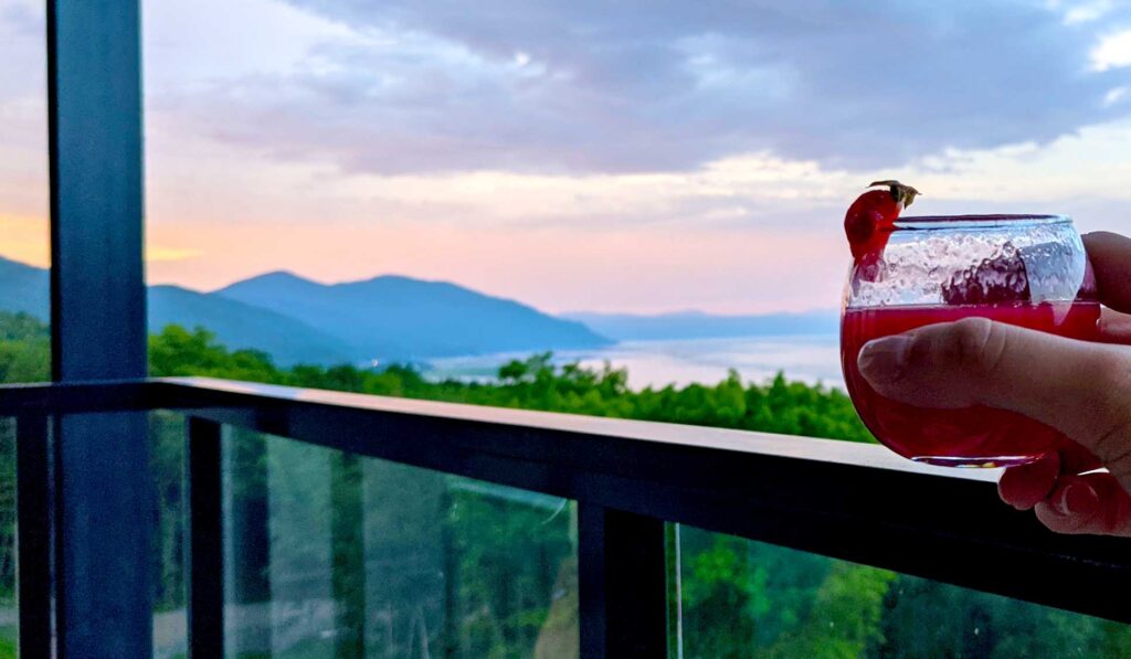 holding-up-a-strawberry-daquari-on-the-balcony-at-club-med-charlevoix-with-the-sunsetting-in-pink-and-blues-over-mountains-and-the-river