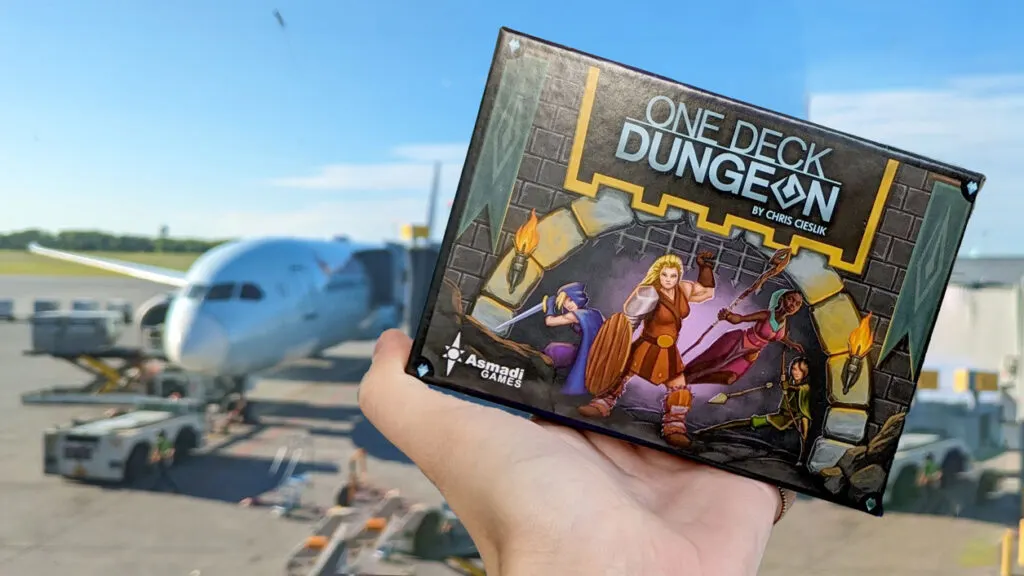 a hand holding up a box that says: One deck dungeon,an arch to a dungeon and 5 women warriors standing in. The box isn't big, fitting in a hand. Behind the hand is a blurred plane connected to the airport by the tunnel