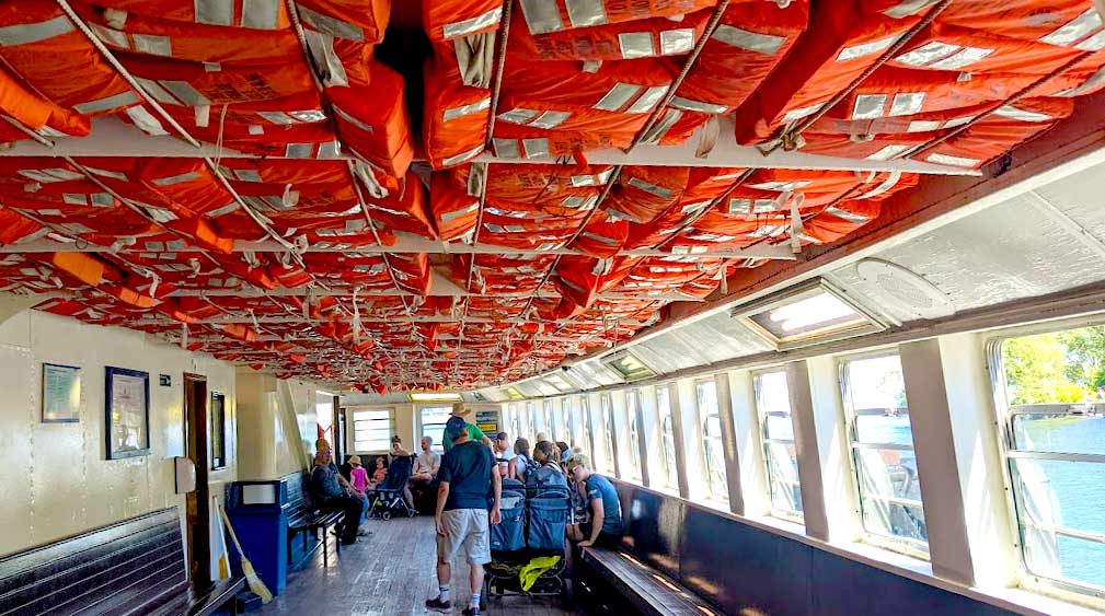 on the toronto island ferry. Benches line the sides of the boat, people are already seated in the back. Above you on the ceiling are thousands of orange life jackets 