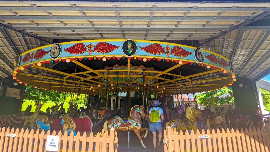 antique carousel on centreville amusement park toronto island. A brown fence separates the carousel, behind it you have a brown horse directly in front. It's pulling a bright red carriage behind it. A woman is walking between the horses and carriages to find her seat. The roof of the carousel is brightly painted with yellow, and blue, with red wings as decor