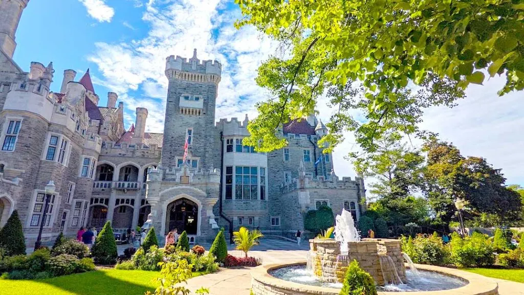 things-to-do-in-toronto: visit casa loma. a medievale castle with a front tower and a beautiful stone water fountain with lush green tree leaves on the edge of the photo