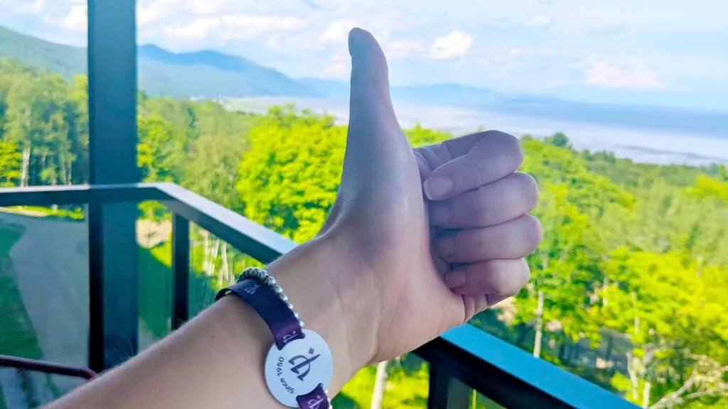 club-med-bracelet-things-to-know
a womans hands giving a thumbs up with wearing a purple ribbon with a club med logo in the middle. shes on a balcony you can see the river, green trees, and mountains blurred in the background on a sunny day