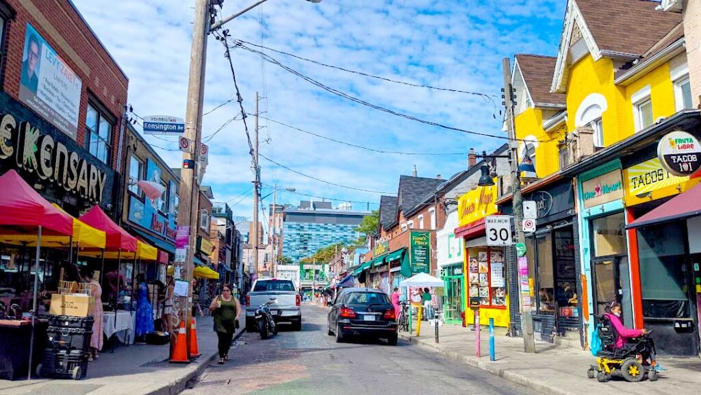 kensington market toronto. Cars are driving down the street with yellow painted buildings on a perfect sunny day.