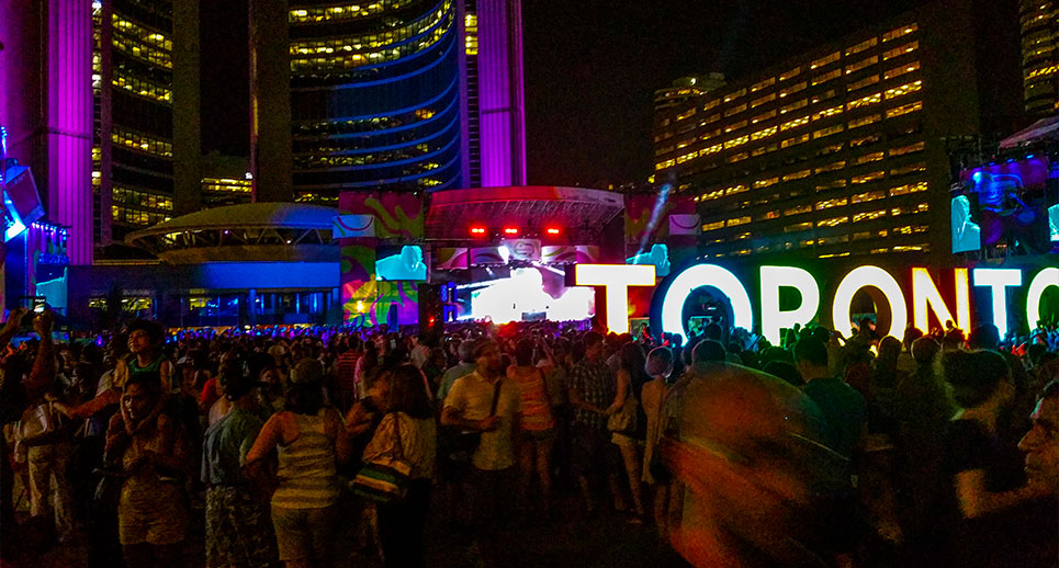 nathan philips square toronto filled with people at night. The toronto sign is lit up in different colours, behind it is a stage with lights so bright you can barely see it.