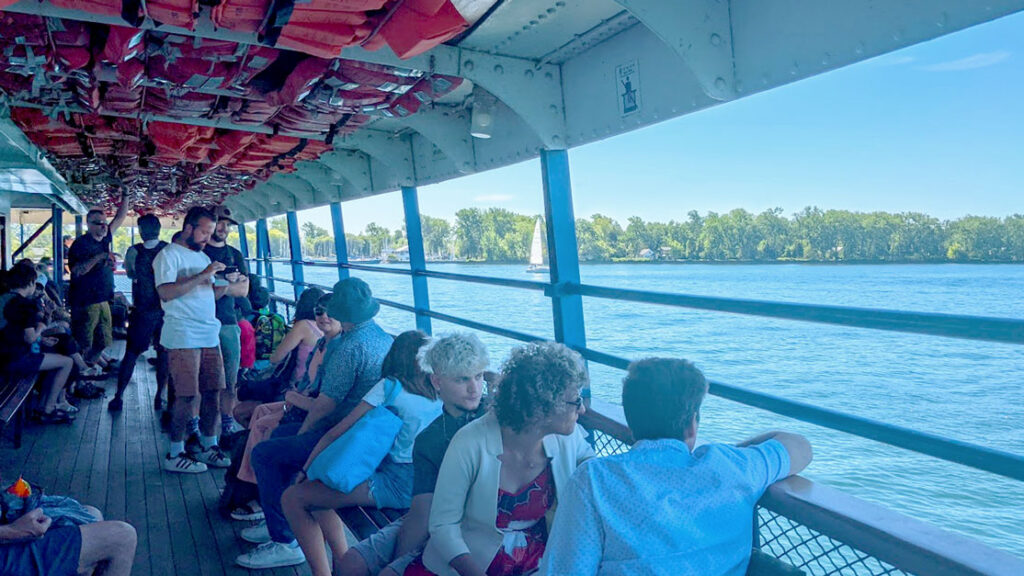 on the upper deck of the toronto island ferry. People are looking out and leaning on the white painted railings to the tree lined island and the water below. On the ceiling of the ferry are bright orange life jackets