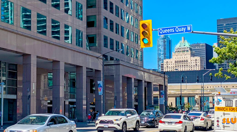 queens quay west toronto. a bustling street in downtown toronto with cars stuck in traffic at a light, despite it being green. On the light post there's a street sign for queens quay w