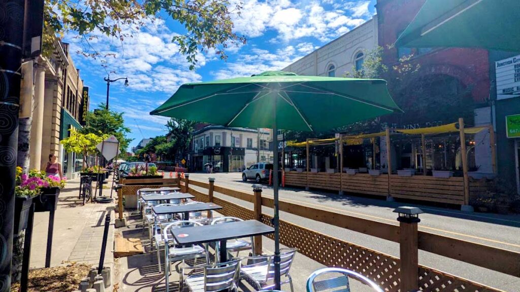the junction toronto. A line of tables and chairs on the street blocked by a brown wooden fence. Green umbrellas are open above them on a bright sunny barely cloudy day. There's  restaurant across the street with the same outdoor terrace on the street. 