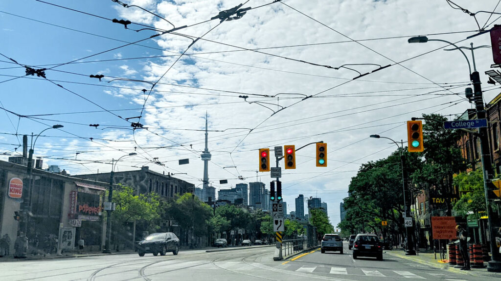 Toronto wine lane road, stopped at a red line. There are streetcar tracks on the street, and the wires line the skyline view. Half the sky is blue while the other half is completley covered in clouds. You can see the CN tower above the buildings lining the road.