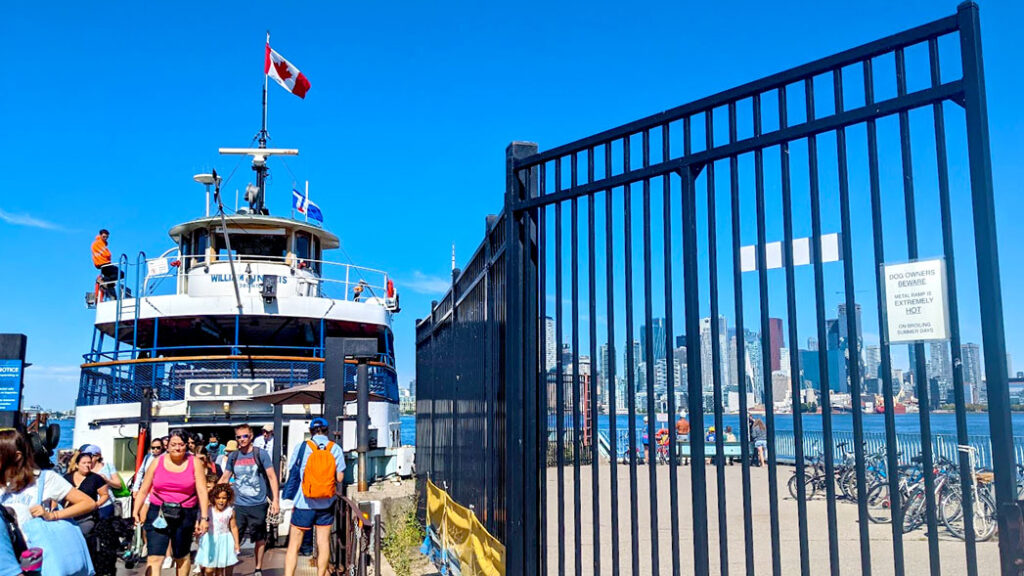 toronto island ferry just got to the dock, people are piling out of the entrance going on to toronto island. there are tall black iron gates blocking your view of the toronto skyline view. Theres a sign on  the ferry that says city. 