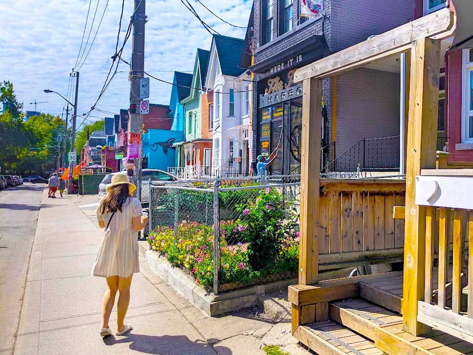a woman in a dress with a wide brimmed sun hat walking on the sidewalks in kensington market in toronto next to brightly painted colorful and tall homes on the side