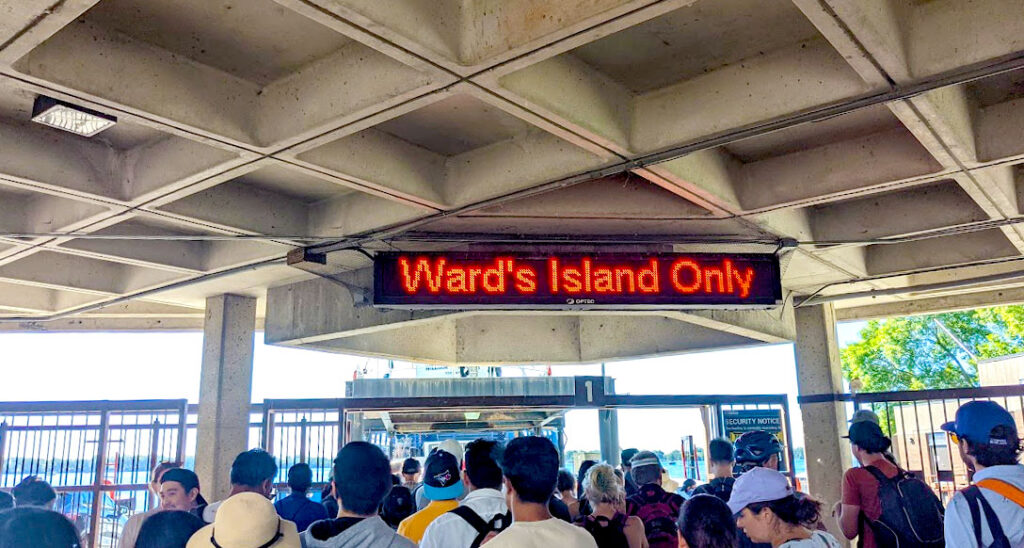standing in line for the toronto island ferry, a lot of people in front, a cement roof above holding up an LED sign saying Ward's Island Only in red. 