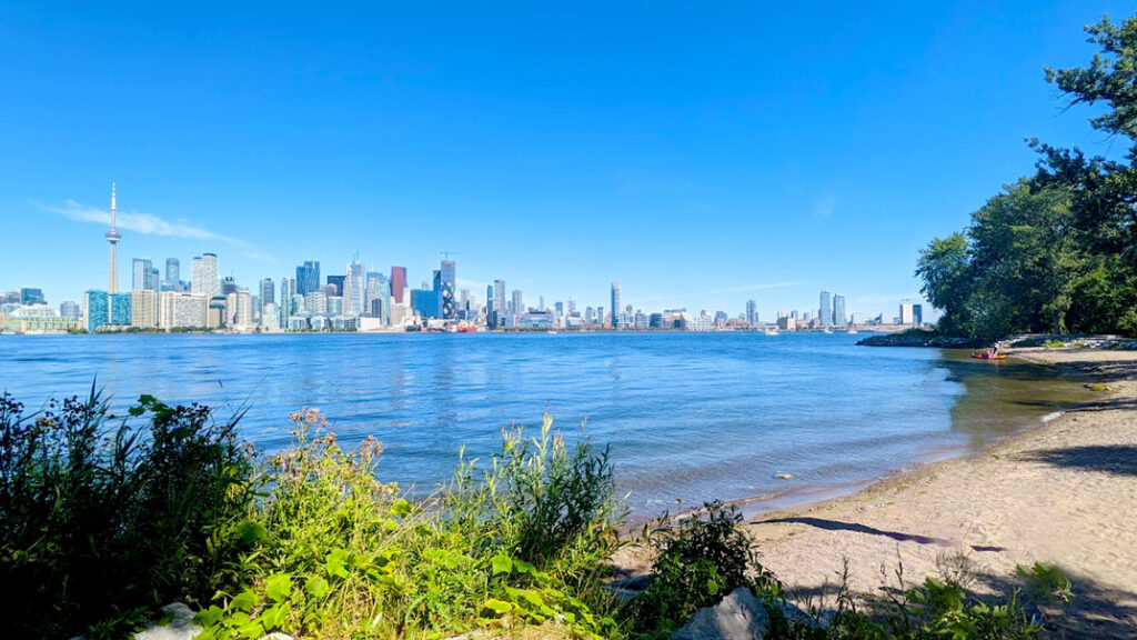 wards island toronto island guide hack: a sandy beach on the shore of ward's island looking out onto the toronto skyline view of all the high rise buildings but the tallest one is the CN tower above them all
