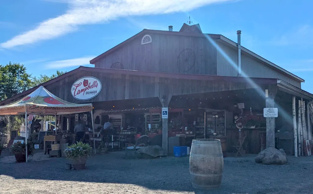 a large woooden building with an overhang covering a fresh produce shop. On the roof is a sign for campbells orchard and a smiling apple logo. This is one of the top things to do in prince edward county