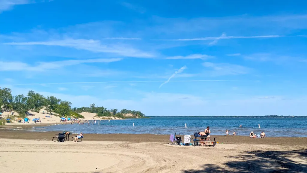 sand dunes provincial park what to do in pec: a flat beach with people set up at picnic tables, while others are enjoying the water. The further down the beach you go, the sand turns into little mountains with some grassy spots and trees.