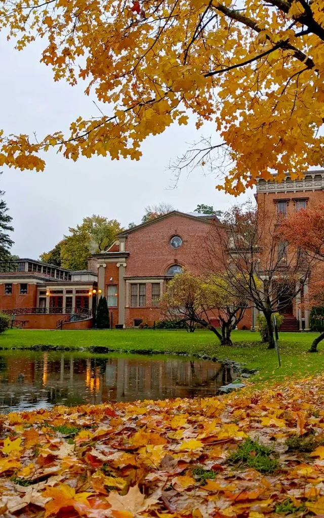 the perfect fall scene in Saratoga Springs. A vertical image. Fallen fall leaves litter the ground from the bottom up until about a third of the way up. Then you can see from grass and a pond, with a slight reflection. Beyond the grass is a red brick building, that goes into a grey sky and finally a tree with yellow and orange leaves breaks into the frame from the right side covering the sky and the top third of the photo. 