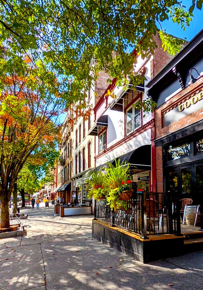 a cozy street in downtown Saratoga springs. Two trees cover the left and top of the photo, the one on top if still green, the blue sky is peaking through. Below it is a small tree and the leaves have started to change colour to orange and yellow. These trees lead you eye to the buildings lining a wide sidewalk. The tallest building is only five stories hight, most are only two. There's often a shop or restaurant on the first floor, two of which have an outdoor patio on the sidewalk. The tree is adding some nice shade to the sidewalk and patios below. The sidewalk is empty, you can slisly make out two figures walking all the way at the end of the sidewalk