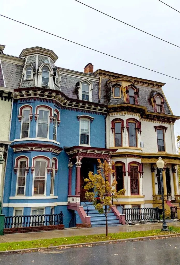 a vertical photo of attached victorian homes in saratoga springs on a rainy day. They are three story homes, with windows in the roof and a bay window on the main floor. You can only see two of the homes, a blue one and a cream white one, but you can see that the blue one is attached to at least one more on the left. It's overcast, but you can tell it had rained before because the street is reflective and much darker than dry pavement