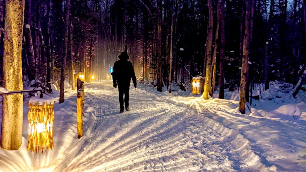 man walking through winter snow path with lanterns lining the forest path