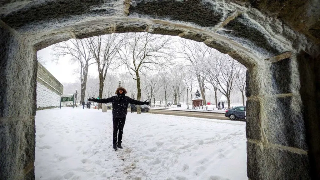 a look through stone wall tunnel. On the other side is a man with outstretched arms. Freshly fallen snow covers the sidewalk. The man is standing in a footpath made by pedestrians as they walk in the snow. trees line the road behind him, they're branches are covered in snow. The man is bundled in a fur line hood as snow is falling in front of his face