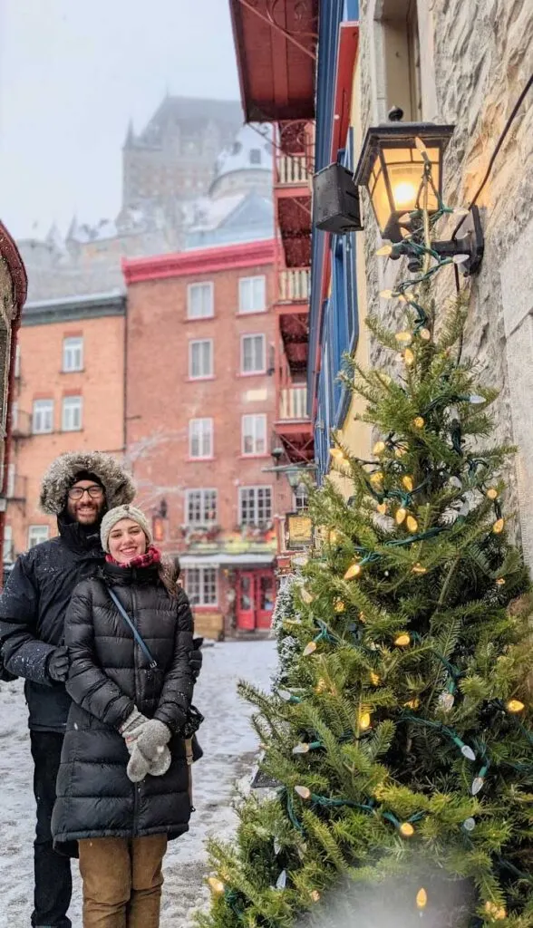 a winter image of quebec city. A woman stands slightly in front of a man, they're both dressed for winter. The mans fur lined hood is over his head for warmth. The woman is wearing a hat and scarf. Both have gloves and winter jackets. They are standing in front of the most iconic view in quebec city: the view of the chateau frontenac from the lower streets in quebec city. There's even a christmas street in front of the couple and a lantern on the side stone wall adding a christmas ambiance to the photo. There's snow on the pavement and it's snowing