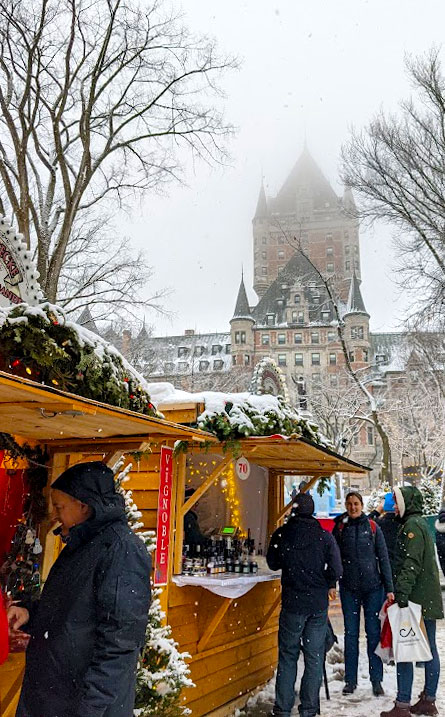 a vertical image. 
A winter scene in Quebec City. 
In the foreground, there are wooden booths with christmas decorations on the front. Consumers are dressed in their biggest winter jackets, looking in at the booths, shopping. Above the booths are bare trees and a completely white sky. But you can still see one of the towers of the Fairmont Chateau Frontenac in the distance