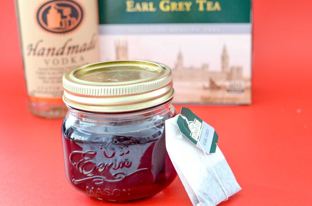 hot-tea-cocktails-vodka-infused-earl-grey-ingredients: a mini mason jar filled with earl grey tea and a tea bag leaning on it . Blurred in the background is a bottle of vodka and the box of earl grey tea. All on a popping red background