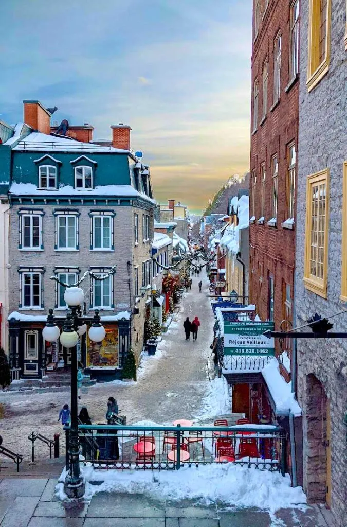 quebec city golden hour. The top of the sky is still blue but once its hits the top of the buildings it starts turning golden yellow. You can see the whole way down a narrow pedestrian only street. The pathway is slushy snow, with white snow on the corners, where people haven't walked. 