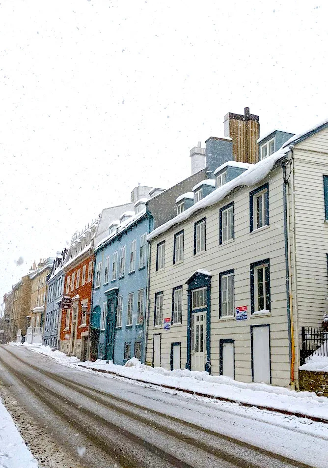 a quiet side street in quebec city in the winter. There's a build up of snow on either side of the road, it's gross slush where the cars drive, revealing the grey pavement below. Homes sit on the right side of the street. There doors are directly on the street in front of the, without a lawn. Despite all the houses being attached, you can tell them apart by the different colours they've been painted. The first one is white, then blue, then burnt orange. The sky is completely white as more snow falls on the street. You can even see some white snow flakes falling in front of the camera. 