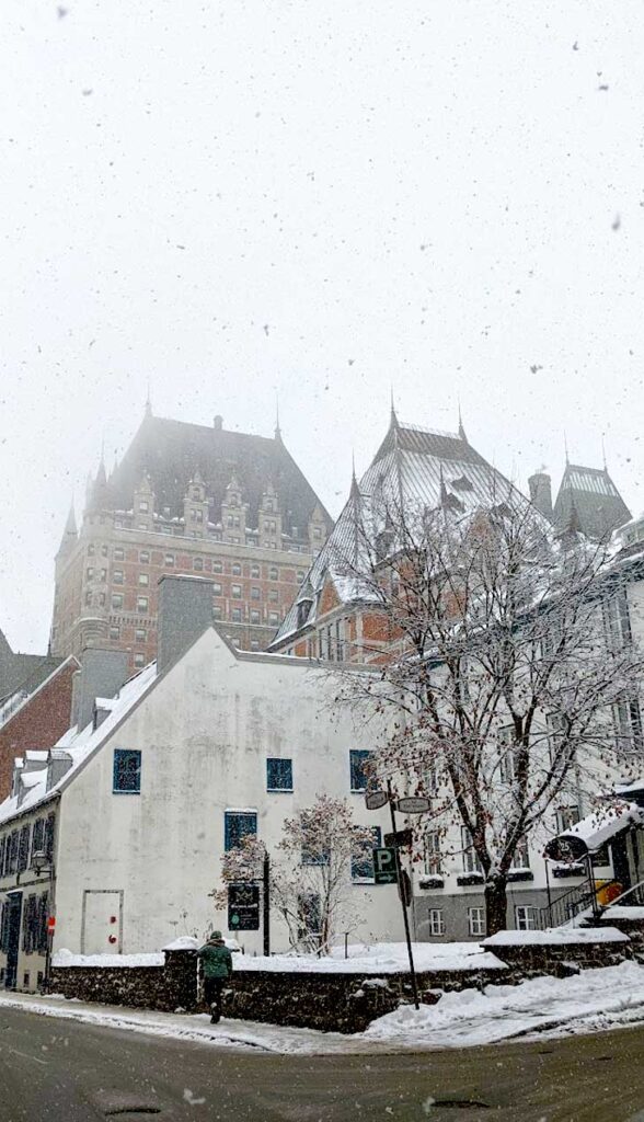 a vertical image of the snowfall in front of the chateau frontenac in quebec city. It's the corner of a street, with an old white, three story building on the corner. Behind the building is the tower of the chateau frontenac, with red bricks and a slanted triangle brown roof. It's almost blended in to the white sky above it as the snow falls.