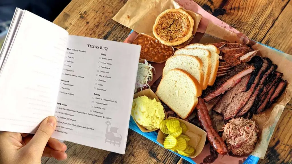 a hand holding a booklet open to a page with the title Texas BBQ it has a checklist of everything you should order from meats, to sides, to desserts, and drinks. With a suggestion of BBQ joints at the bottom. In the background you see a food tray filled iwth meats and sides from a Texas BBQ