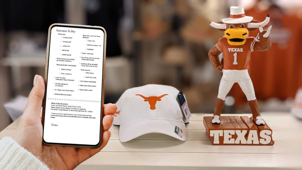 a hand holding a phone showing the screen to the camera. On the screen is a souvenir checklist. Behind her, is a white and orange texas longhorn hat and figurine - great texas souvneirs. 