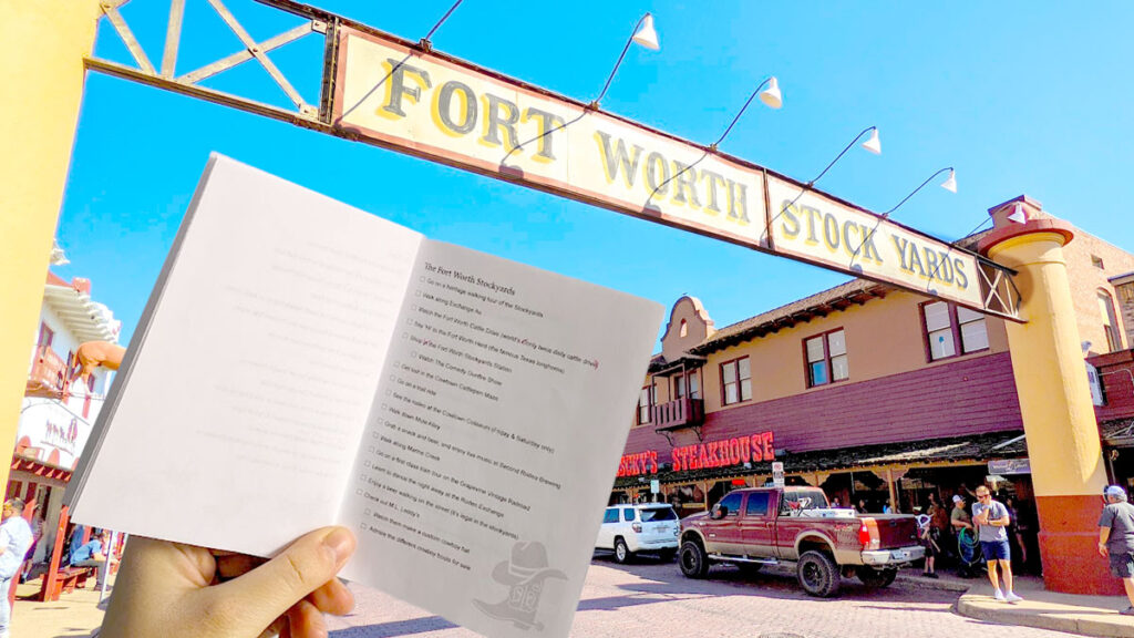 a hand holding a small pocket size printed booklet with a checklist for the fort worth stockyards. In the background is the opening sign for the stockyards that goas above the road, connecting both sidewalks. 