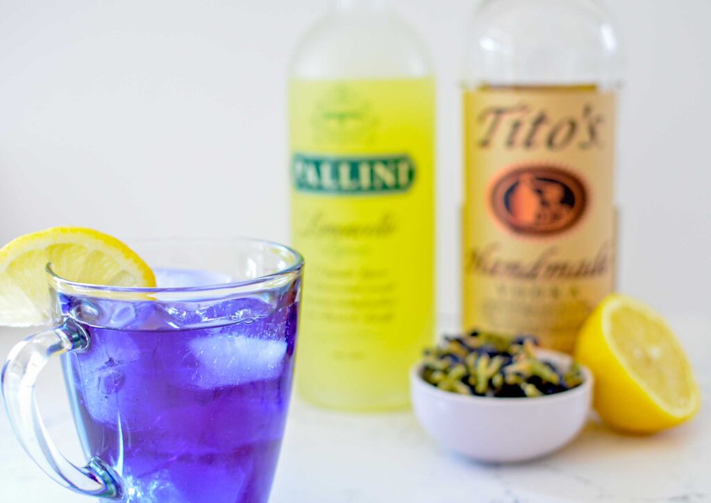 iced-butterfly-pea-flower-tea-cocktail in the front with a lemon wedge on the glass rim. Blurred in the background is the bottle of limoncello, titos vodka, a bowl with butterfly pea flower petals, and a half a lemon