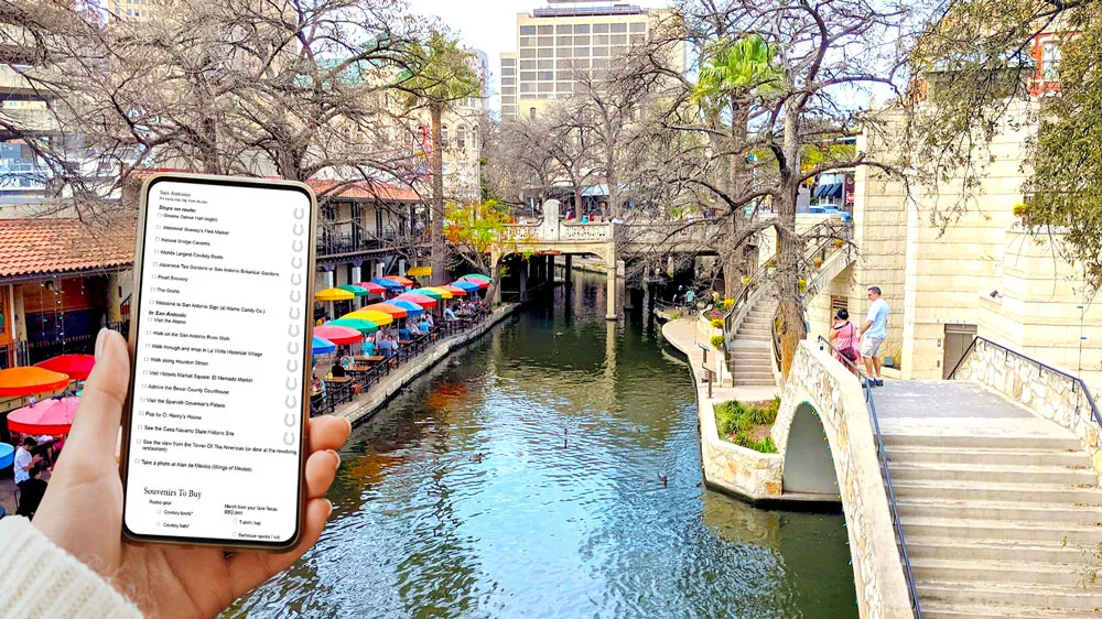 a hand holding a cellphone open to a document with a san antonio checklist being displayed. In the background is the san antonio river walk with an arched bridge connecting two sides. On the sidewalk on the left it's lined rainbow colour umbrellas covering tables at a restaurant. 