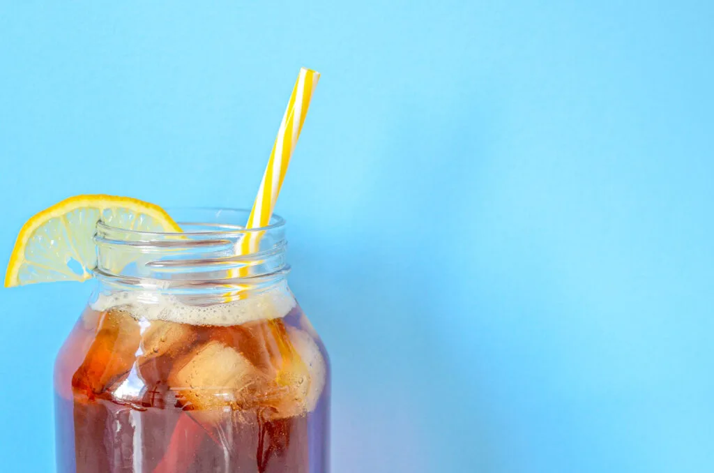 iced-tea-with-lemon-slice in a large mason jar against a blue background and a yellow striped straw in the glass