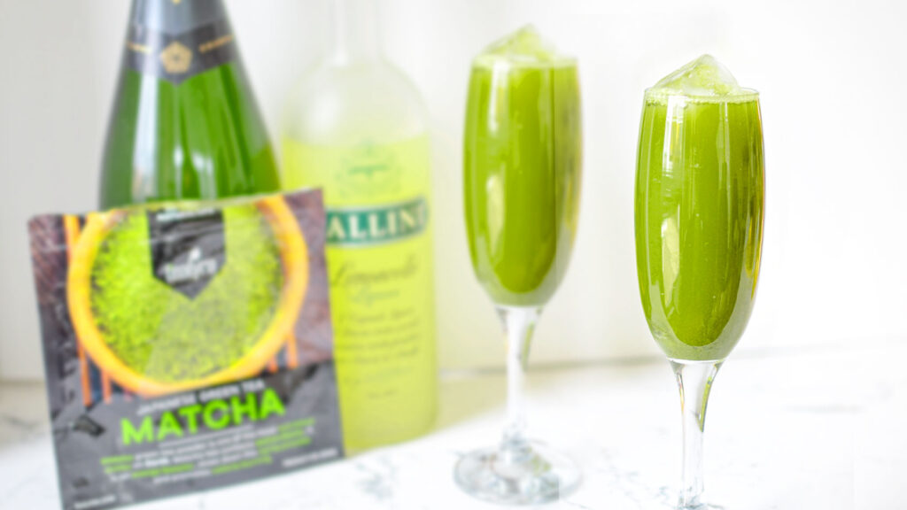 2 matcha-spritz-green-tea-cocktails in champagne glasses with floating ice cubes and blurred in the background a matcha package, champagne bottle, and the bottle of limoncello